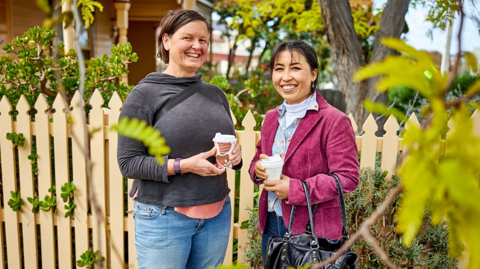 Two female Diversitat team members, posing for photo in garden. Cream picket fence is seen in background.  