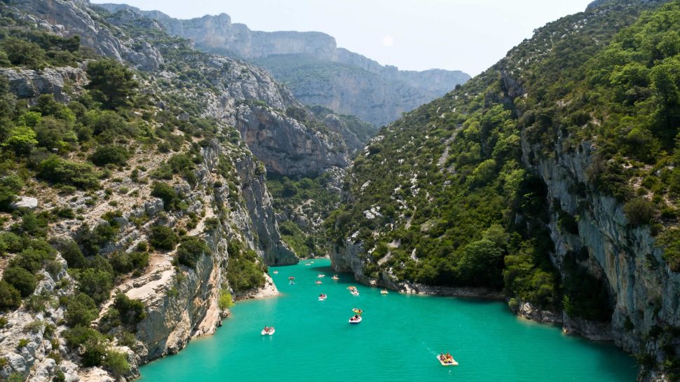 A panoramic view of the Verdon Gorge in France. Azure, still water runs through the middle of this scene, subtly winding through the rock formations that surround it. Huge rocks forming the gorge stand tall to both the left and right. There’s dense shrubbery forming on the rocks. People can be seen kayaking through the crystal blue water. 